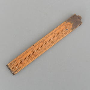 Könnte beinhalten: Ein alter, zusammenklappbarer Holz-Zollstock mit Zentimeter-Messungen von 25 bis 48 cm. Der Zollstock besteht aus hellbraunem Holz mit dunkelbraunen Metallgelenken und Markierungen. Der Zollstock ist leicht geöffnet.