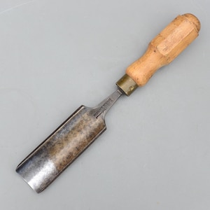 May include: A vintage woodworking chisel. It features a wooden handle and a metal blade. The blade has a slightly curved shape and a worn appearance. The handle is light brown and the metal blade is a darker, aged colour.