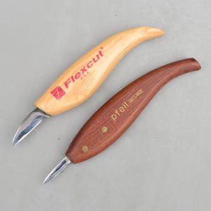 May include: Two wood carving knives. One is light-colored with "Flexcut USA" printed in red. The other is dark brown with "pfeil SWISS MADE" printed in gold. Both knives have curved blades and ergonomic handles.