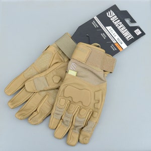 May include: A pair of tan leather tactical gloves with a velcro wrist strap and knuckle protection. The gloves are labelled "BLACKHAWK! S.O.L.A.G. RECON" and feature touch screen compatible fingertips.