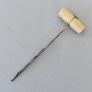 May include: An antique awl. It has a cream-coloured handle and a metal shaft. The handle is cylindrical and appears to be made of bone or ivory. The metal shaft tapers to a sharp point. The awl is likely used for piercing materials.