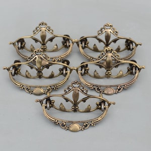 May include: Five antique brass-toned metal drawer pulls arranged in a group. Each pull features a decorative, ornate backplate with scrollwork and a central vertical element, connected to a curved, swinging handle with a smooth, rounded bottom edge.