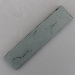 May include: A rectangular, gray sharpening stone with a textured surface and dark crack-like markings. The stone is angled, showcasing its length and the subtle variations in its color and texture.
