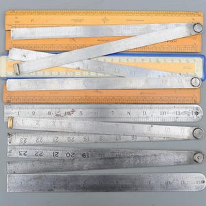 Puede incluir: Una colección de reglas antiguas, incluyendo tipos plegables y rectos. Las reglas son de madera y metal, con marcas de medición en centímetros. Algunas reglas son de color naranja, azul y plateado.