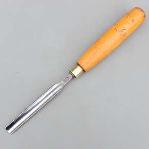 Peut inclure: Un ciseau à bois avec un manche en bois et une lame en métal. Le manche est brun clair et la lame est argentée. Le ciseau est conçu pour sculpter et façonner le bois. L'outil est incliné et a un bord tranchant.