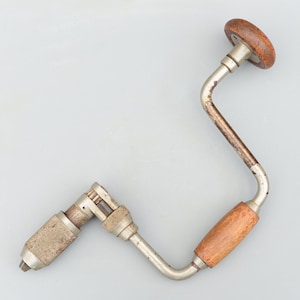 Peut inclure: Une perceuse à main vintage avec une poignée en bois et une manivelle en métal. La perceuse est argentée avec de la rouille et a une poignée en bois.