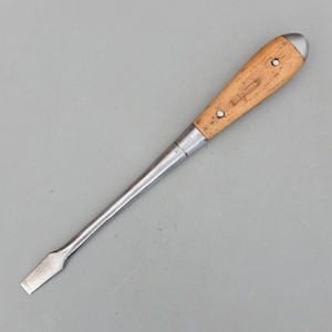 Può includere: Un cacciavite a taglio vintage con manico in legno e lama in metallo. L'impugnatura è marrone chiaro con un cappuccio in metallo e due rivetti. La lama è argentata e rettangolare. L'utensile è progettato per avvitare.