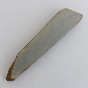 以下が含まれることがあります： わずかに角度の付いた形状の灰色の砥石。石は長方形で、片方の端に茶色でざらざらしたエッジがあります。石の表面はくすんだ灰色で、工具の研ぎに使用されていることを示唆しています。