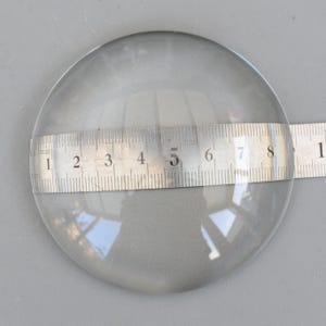 May include: A clear, round glass lens with a ruler laid across it. The lens appears to be approximately 3 inches in diameter. The ruler is marked in inches and millimeters.