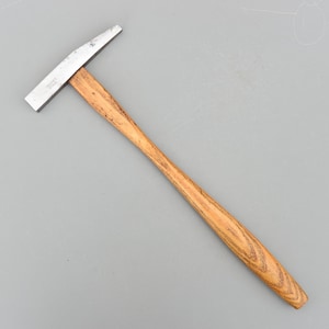 Può includere: Un martello con testa in metallo e manico in legno. La testa del martello è argentata e il manico è marrone chiaro con venature del legno. L'utensile è progettato per lavori di costruzione o riparazione. Il manico misura circa 25 cm.