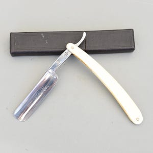 May include: A straight razor with a cream-coloured handle and a silver blade, open and ready for use. The blade is marked with the text "BELOW GROUND". A black case is in the background.