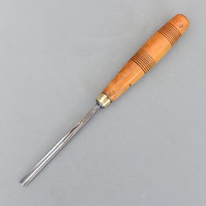 May include: A wood chisel with a light brown handle and a silver blade. The handle has horizontal grooves and a brass ferrule. The blade is marked with text. A woodworking tool.
