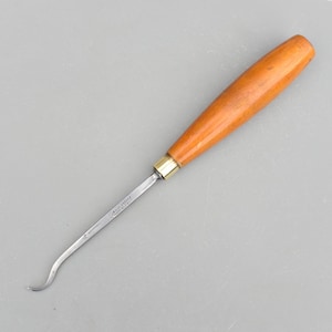 May include: A wood-handled carving tool with a curved blade. The handle is a light brown color, and the blade is silver. The tool has a brass ferrule where the blade meets the handle. The tool is designed for detailed carving work.