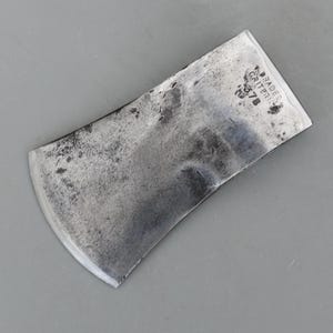 Puede incluir: Una cabeza de hacha vintage con una hoja curva y una parte superior rectangular. El metal tiene un aspecto desgastado y texturizado con manchas oscuras. El texto "BRADES CRITERION 378" está estampado en la parte superior.