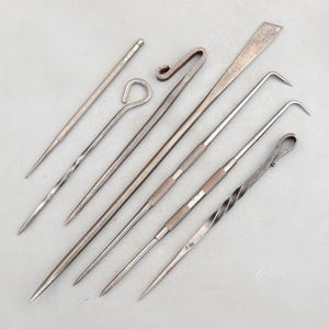 May include: Collection of antique metal tools, including picks, hooks, and a lever. The tools vary in shape and size, with pointed ends and textured handles. The tools are arranged on a light gray surface.