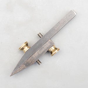 May include: A vintage metal marking gauge with a pointed end and brass accents. The gauge has a screw for adjustment and the text "MOUNTFORD" is visible. The tool is designed for woodworking or similar crafts.