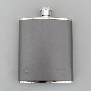 May include: A rectangular, gray flask with a silver rim and cap. The flask has the words "British Airways" printed on the front. The cap is silver and has a ridged design.