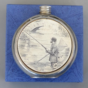May include: A silver-toned flask with a circular, cream-colored face featuring a black and white illustration of a man fishing. The flask is set against a blue textured background. The initials "H.H." are visible.