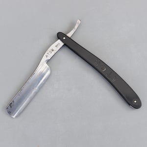 May include: A vintage straight razor with a silver blade and a black handle. The blade is marked with the text "ATOR REG 9". The handle is curved and features the word "ATOR". The razor is open, displaying the sharp blade.
