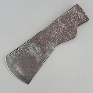 May include: A hand-forged metal axe head featuring a dark, textured, hammered surface with a wide, curved cutting edge. The metal displays a raw, uneven finish with visible indentations across the body, tapering toward a narrow, flat tang.
