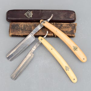 Puede incluir: Dos navajas de afeitar antiguas con mangos de marfil y una caja de madera. Las navajas tienen la inscripción "TRES BIEN TEMPRE" y un logotipo de diamante. La caja tiene la inscripción "SHEFFIELD" y un logotipo de diamante.