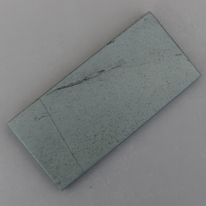May include: A rectangular, gray sharpening stone with a speckled surface. The stone has a slightly rough texture and a dark line running across its surface. The stone is designed for sharpening knives and other tools.