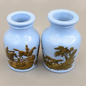Peut inclure: Deux vases en céramique bleu clair avec un motif en relief doré. Le motif représente des scènes équestres avec des cavaliers à cheval et des chiens de chasse. Les vases ont un corps arrondi et un bord évasé.
