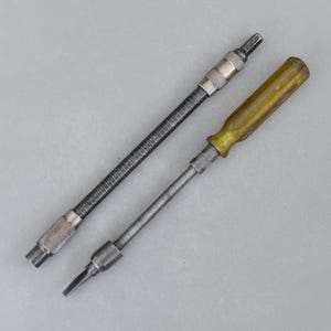 Puede incluir: Dos destornilladores flexibles. Uno tiene un eje negro y corrugado con extremos de metal plateado. El otro tiene un mango amarillento y transparente y un eje de metal plateado. Ambas herramientas están diseñadas para alcanzar ángulos difíciles.