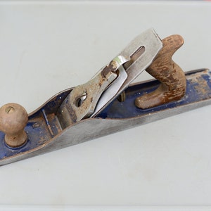 Può includere: Un pialla a mano vintage blu e argento con manico in legno. La pialla ha una lama in metallo e una manopola sul davanti.