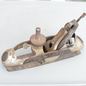 Op de afbeelding: Een antieke Stanley No. 20 blokschaaf, met een metalen behuizing en zichtbare roest. De schaaf heeft een mes, een stelschroef en een hendel. Deze houtbewerkingsgereedschap is ontworpen voor het gladmaken van oppervlakken.