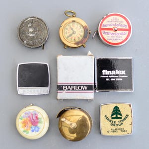 Puede incluir: Surtido de cintas métricas vintage, que incluyen una pequeña cinta métrica de estilo reloj de bolsillo, una cinta métrica redonda con un diseño floral, una cinta métrica cuadrada con el texto "Parker Timber Group" y un número de teléfono, y una cinta métrica rectangular con el texto "finalex" y un número de teléfono.