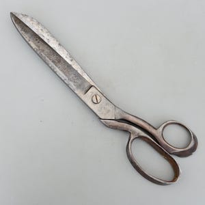 May include: A pair of vintage metal scissors with a worn, aged appearance. The blades are long and pointed, with a visible screw at the pivot point. The handles feature finger holes for grip. The scissors are likely used for cutting fabric or paper.