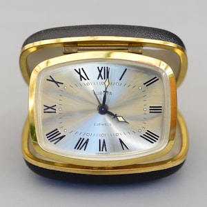 May include: A vintage Europa travel clock in a black and gold case. The clock face is silver with black Roman numerals and hands. The words "EUROPA" and "2 JEWELS" are printed on the face. The case is rectangular with rounded corners.