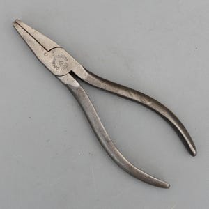 May include: A pair of vintage metal needle-nose pliers with a dark, weathered finish. The tool features long, tapered jaws with a serrated gripping surface and curved handles. The pivot point is stamped with the text Grumpstone Co 1955.