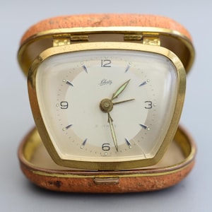 May include: A vintage Schatz travel clock in an open, orange leatherette case. The clock has a gold-tone bezel and a cream-coloured face with black numerals and hands. The clock face reads "Schatz" and "2 Jewels".