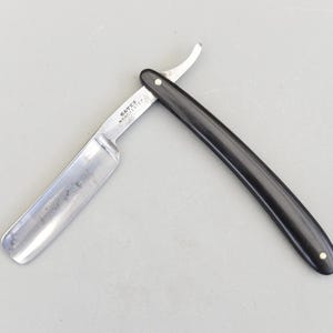 May include: A straight razor with a polished silver blade and a black handle. The blade is open, showing the cutting edge. The handle is curved and has a small stud at the end. The text "BATES MANCHESTER" is visible on the blade.