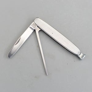 May include: A silver pocket knife with a textured handle and a pointed tool. The knife blade is open, revealing the sharp edge. The handle has a diamond pattern. The tools are designed for pipe smoking.