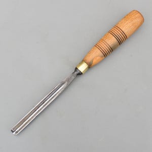 Könnte beinhalten: Ein Holzbeitel mit einem hellbraunen Holzgriff, der mit dunkelbraunen Ringen verziert ist, und einer silbernen Metallklinge. Das Werkzeug ist für die Holzbearbeitung und das Schnitzen konzipiert. Die Klinge ist aus Metall.