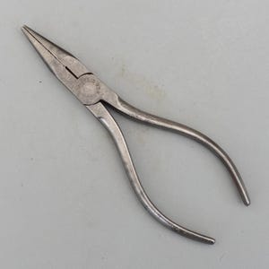 May include: A pair of needle-nose pliers made of silver-colored metal. The pliers have long, tapered jaws and curved handles. The tool is designed for gripping, bending, and cutting wires and other small objects. The pliers are lying on a gray surface.