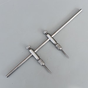 May include: Two silver adjustable dividers with pointed tips and knurled knobs, lying on a grey surface. The arms of the dividers form a cross shape. These appear to be precision measuring instruments, likely used in engineering or drafting.