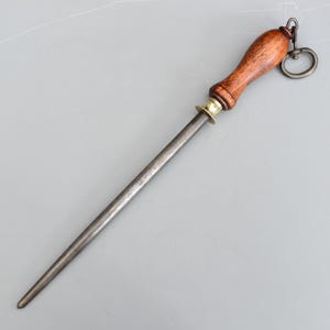 May include: A knife sharpening steel with a wooden handle and a metal ring for hanging. The steel is cylindrical and tapers to a point. The handle is a light brown colour, and the metal is a dark grey.