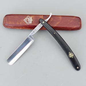 May include: A straight razor with a silver blade and a black handle, resting on a wooden case. The blade is marked with the brand "PUMA" and "SOLINGEN". The case is brown with the Puma logo.