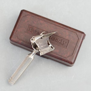 Puede incluir: Una navaja Ever-Ready vintage de color plateado, con un mango texturizado y una hoja plegable. La navaja está en una caja de plástico marrón con las palabras "Ever-Ready" en relieve en la parte superior.