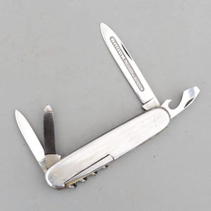 May include: A silver-toned multi-tool pocket knife with a brushed metal handle. The tool features an extended main blade engraved with the text BRÜCK PRESSEN U. SCHEREN, a smaller blade, a nail file, and a bottle opener integrated into the handle.