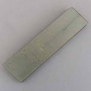 以下が含まれることがあります： 長方形の、薄緑色の砥石で、表面は少しざらざらしています。石の表面には、対角線上に走るひび割れが見えます。このオブジェクトは、刃物や工具を研ぐために設計されています。