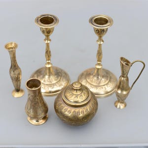 May include: A collection of antique brass decorative items, including two candlesticks, two vases, a small pitcher, and a lidded container. The items have an ornate, engraved leaf design and a warm, golden hue.