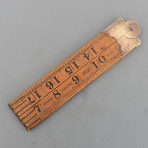 May include: A vintage wooden folding ruler with brass fittings. The ruler is marked with black numerals and lines, and the text "RABONE" and "MADE IN ENGLAND" are visible. The ruler measures up to 43 cm.