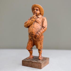May include: A wooden carving of a man wearing a hat and a long-sleeved shirt. He is standing with his hands clasped in front of him, and he has a thoughtful expression on his face.