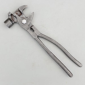 May include: A multi-functional hand tool made of dark grey metal, combining a hammer head on one side with adjustable pliers featuring serrated jaws and a central hexagonal bolt pivot point, designed for gripping, turning, and striking tasks.