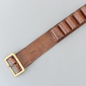 May include: A brown leather cartridge belt with loops for holding ammunition. It has a brass buckle and is designed to be worn around the waist. The leather has a worn appearance, suggesting age and use. Likely for hunting or shooting sports.